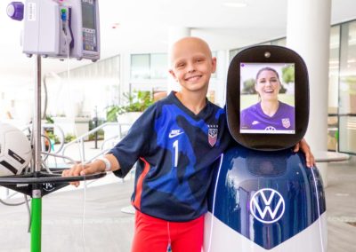 Young fan Luna Perrone, at the hospital, smiling with her arm around CHAMP as she visits remotely with USWNT forward Alex Morgan